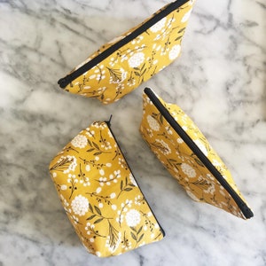 May include: Three yellow fabric pouches with black zippers and a white floral pattern. The pouches are on a white marble surface.