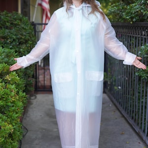 May include: A clear, hooded raincoat with a button-up front and two front pockets. The long, transparent coat is worn over a light blue garment. The person is wearing gold-colored heels.