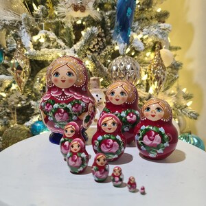 May include: A set of traditional Russian nesting dolls, or Matryoshka dolls, in varying sizes. The dolls are primarily red with floral designs and painted faces. The dolls are arranged on a white surface with a decorated Christmas tree in the background.