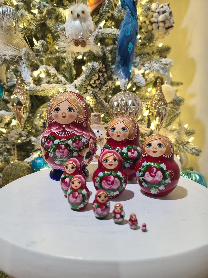 May include: A set of traditional Russian nesting dolls, or Matryoshka dolls, in varying sizes. The dolls are primarily red with floral designs and painted faces. The dolls are arranged on a white surface with a decorated Christmas tree in the background.