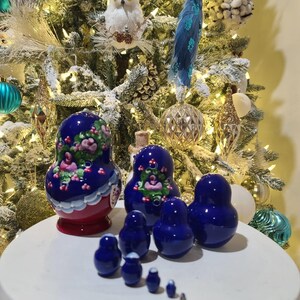 May include: A set of Russian nesting dolls in varying sizes. The largest doll is painted with a floral design in pink and white on a blue background. The smaller dolls are solid blue. The dolls are displayed on a white surface in front of a decorated Christmas tree.