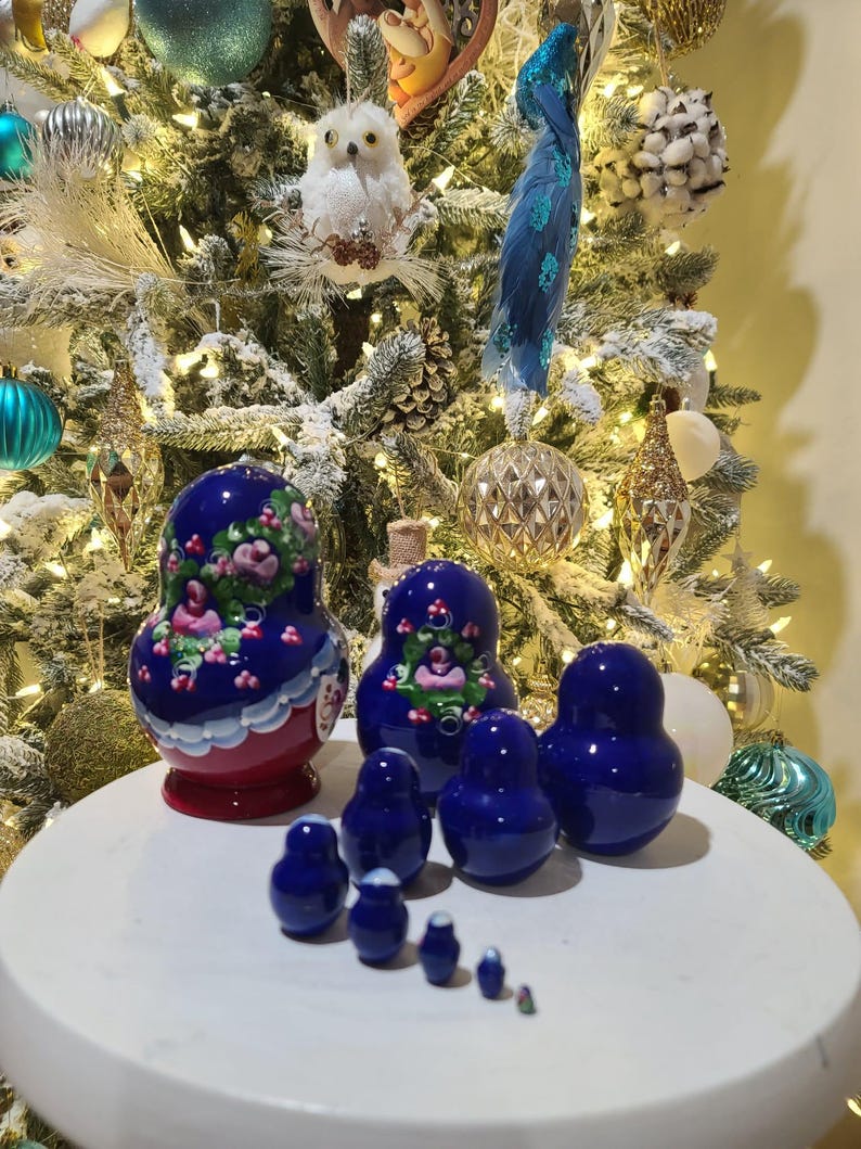 May include: A set of Russian nesting dolls in varying sizes. The largest doll is painted with a floral design in pink and white on a blue background. The smaller dolls are solid blue. The dolls are displayed on a white surface in front of a decorated Christmas tree.