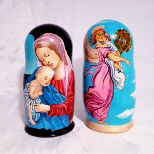 May include: Two hand-painted wooden nesting dolls, or matryoshka dolls. The larger doll depicts the Virgin Mary holding the baby Jesus. The smaller doll depicts an angel holding a child. Both dolls have a blue background with clouds.