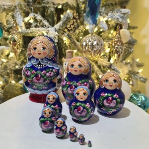 May include: A collection of Russian nesting dolls, or Matryoshka dolls, in various sizes. The dolls are painted with blue, pink, and white floral designs and feature smiling faces. The dolls are arranged on a white surface, with a decorated Christmas tree in the background.
