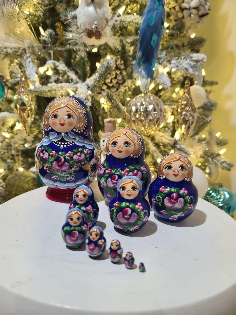 May include: A collection of Russian nesting dolls, or Matryoshka dolls, in various sizes. The dolls are painted with blue, pink, and white floral designs and feature smiling faces. The dolls are arranged on a white surface, with a decorated Christmas tree in the background.