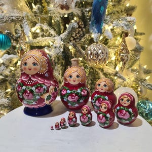 May include: A set of traditional Russian nesting dolls, or Matryoshka dolls, in varying sizes. The dolls are painted red with floral designs and feature a smiling woman's face. The dolls are displayed on a white surface with a decorated Christmas tree in the background.