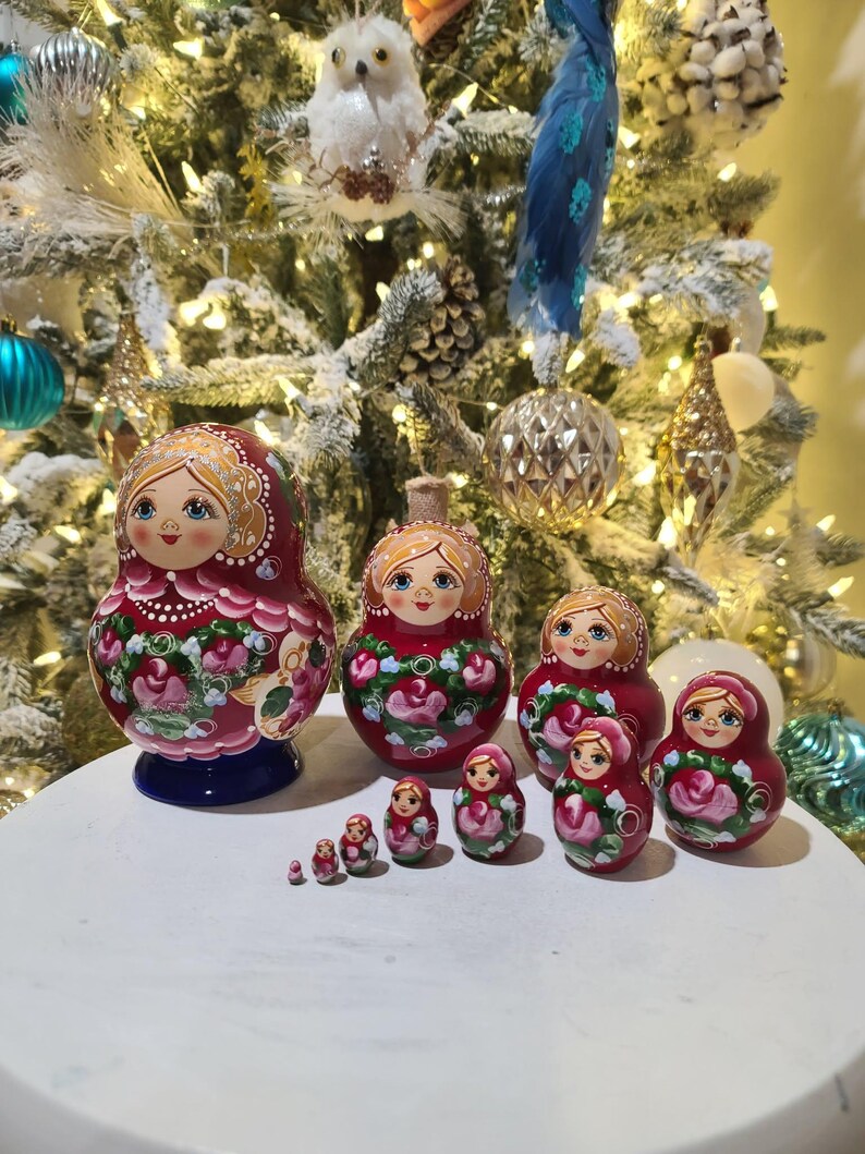 May include: A set of traditional Russian nesting dolls, or Matryoshka dolls, in varying sizes. The dolls are painted red with floral designs and feature a smiling woman's face. The dolls are displayed on a white surface with a decorated Christmas tree in the background.