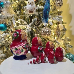 May include: A set of red nesting dolls with floral and owl designs. The dolls vary in size, with the largest doll having a blue base. The dolls are arranged on a white surface in front of a decorated Christmas tree.