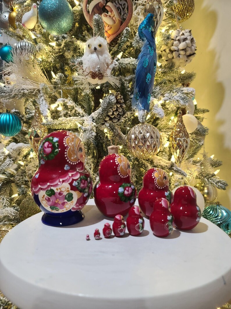 May include: A set of red nesting dolls with floral and owl designs. The dolls vary in size, with the largest doll having a blue base. The dolls are arranged on a white surface in front of a decorated Christmas tree.