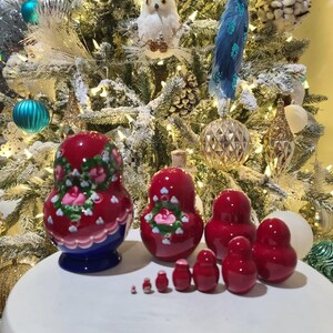 May include: A set of red nesting dolls, also known as Matryoshka dolls, with floral designs. The largest doll has a blue base. The dolls are arranged on a white surface in front of a decorated Christmas tree.