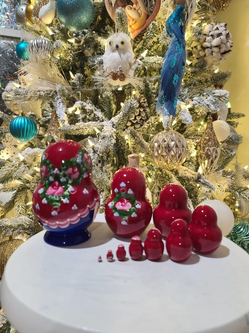 May include: A set of red nesting dolls, also known as Matryoshka dolls, with floral designs. The largest doll has a blue base. The dolls are arranged on a white surface in front of a decorated Christmas tree.