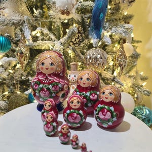 May include: A collection of Russian nesting dolls, or Matryoshka dolls, in varying sizes. The dolls are painted with a red background and floral designs. The dolls are arranged on a white surface, with a decorated Christmas tree in the background.