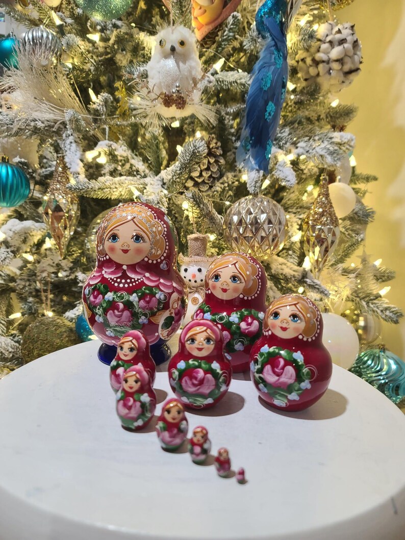 May include: A collection of Russian nesting dolls, or Matryoshka dolls, in varying sizes. The dolls are painted with a red background and floral designs. The dolls are arranged on a white surface, with a decorated Christmas tree in the background.