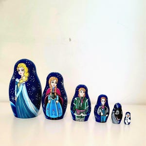 May include: A set of six wooden nesting dolls painted in a dark blue theme with white star accents. The dolls feature hand-painted characters including a blonde woman in a blue gown, a woman with braids, a man in green, a man in a suit, a reindeer, and a snowman.