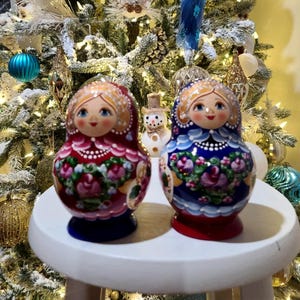 May include: Two Russian nesting dolls, one red and one blue, sit on a white surface. The dolls have painted faces and floral designs. A snowman figurine is between them. The background is a decorated Christmas tree.