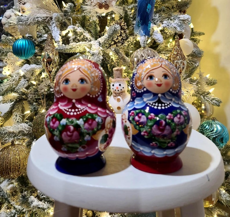 May include: Two Russian nesting dolls, one red and one blue, sit on a white surface. The dolls have painted faces and floral designs. A snowman figurine is between them. The background is a decorated Christmas tree.