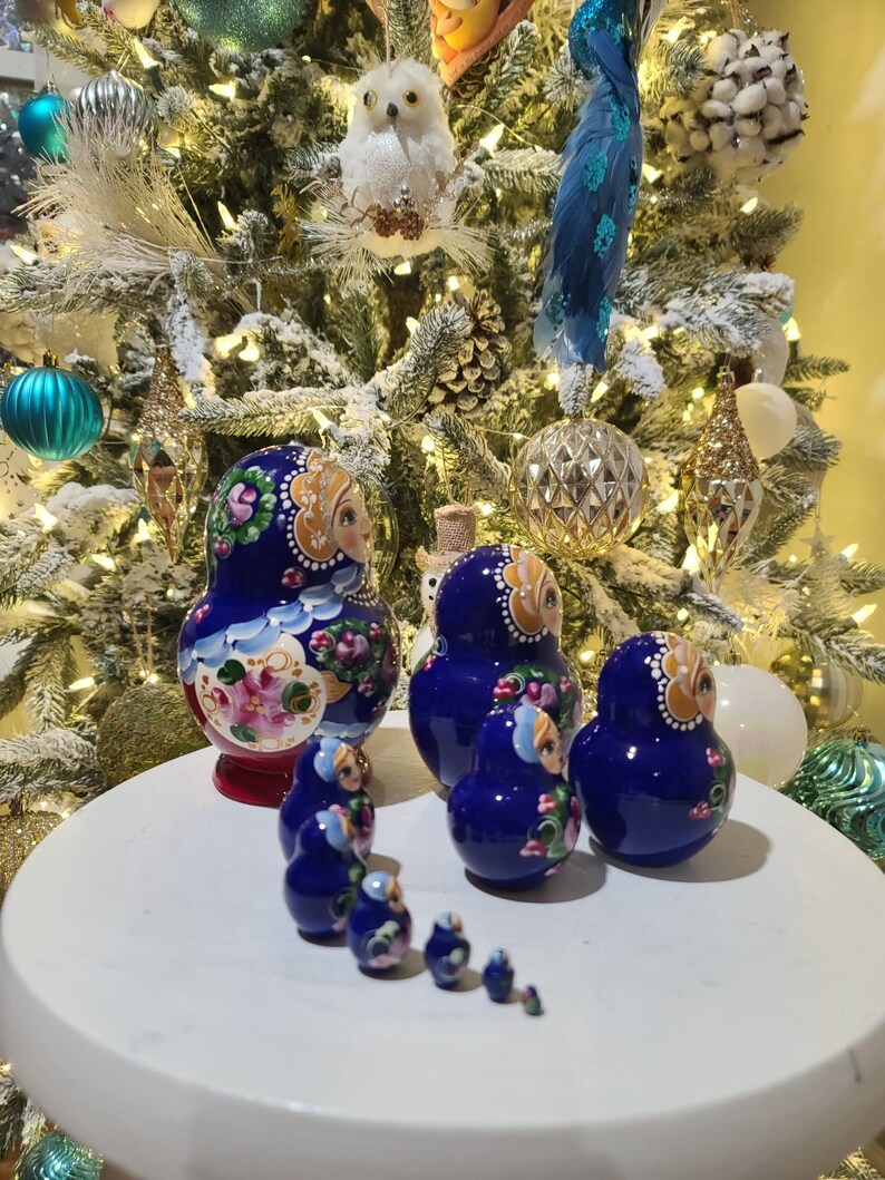 May include: A set of Russian nesting dolls, or Matryoshka dolls, in various sizes. The dolls are painted in a deep blue with floral and face details. The dolls are arranged on a white surface in front of a decorated Christmas tree.
