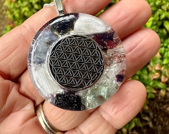 May include: A round pendant with a silver chain. The pendant features a central black metal design surrounded by clear resin and colorful stones. The stones include white, purple, and black. The pendant is held in a hand.