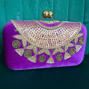 May include: A rectangular, purple velvet clutch with gold embellishments. The top flap features a fan-shaped design of gold sequins and embroidered gold thread. The clasp is two gold spheres. The clutch is a formal accessory.