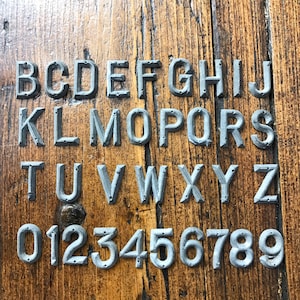 Peut inclure: Un ensemble complet de l'alphabet et des chiffres dans une police grise de style industriel. Les lettres et les chiffres sont montés sur une surface en bois rustique. Chaque caractère a un aspect texturé avec de petits détails en forme de rivets.