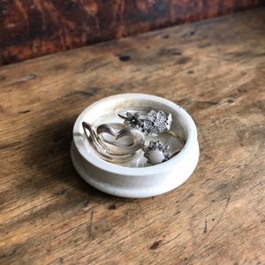 May include: A small, round, off-white ceramic ring dish with a crackle glaze. The dish holds several silver rings, including a heart-shaped ring, a flower ring, and a ring with a central stone. The dish sits on a rustic wooden surface.