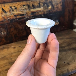 May include: A small, white ceramic cup with a flared rim. The cup has a cylindrical body and a slightly rounded base. The rim shows signs of age. The cup is held in a hand against a blurred wooden background.
