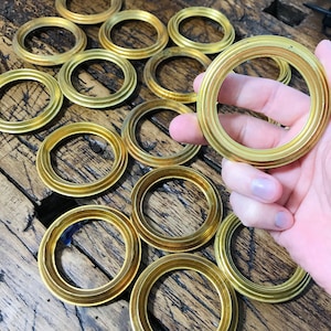 Puede incluir: Anillos de latón dorados con diseño acanalado, dispuestos sobre una superficie de madera. Un anillo es sostenido en la mano, mostrando los detalles del producto. Ideales para proyectos de artesanía o industriales.