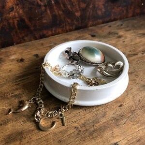 Puede incluir: Un plato de cerámica blanco contiene varias joyas. Una pulsera de cadena dorada con dijes, un anillo de plata con detalles florales y un colgante con una piedra verde son visibles. Los objetos descansan sobre una superficie de madera.