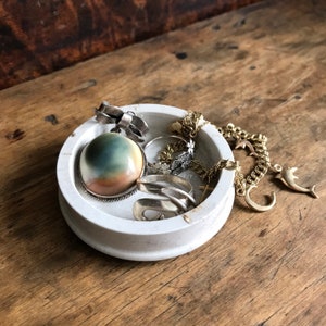 Puede incluir: Un cuenco blanco y redondo para joyas lleno de varias piezas de joyería. Incluye un colgante con un lazo, un colgante redondo grande con una piedra azul y beige, anillos y una cadena dorada con un dije de delfín. El cuenco está sobre una superficie de madera.