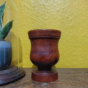 Könnte beinhalten: Eine gedrechselte Holzschale mit einem Sockel. Die Schale hat einen abgerundeten Körper und einen ausgestellten Rand. Das Holz hat eine satte, rotbraune Farbe mit sichtbarer Maserung. Die Schale wird auf einer Holzoberfläche vor einer gelben Wand präsentiert.