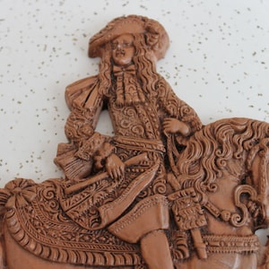 May include: A brown ceramic wall sculpture of a man riding a horse. The man is wearing a hat and a detailed, ornate outfit. The horse has a flowing mane and tail.