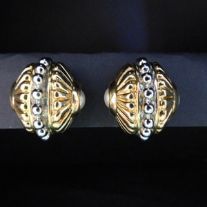 May include: A pair of gold and silver tone earrings with a round, ornate design. The earrings have a textured surface and a pearl-like accent.