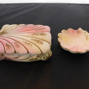 May include: A set of two decorative ceramic seashell trinket boxes. The larger box is closed and the smaller box is open. Both boxes are painted in shades of pink, white, and gold.