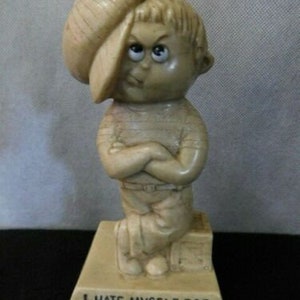 May include: A beige ceramic figurine of a boy with a hat, arms crossed, and a sad expression. The figurine is on a base with the text "I hate myself for hurting you".
