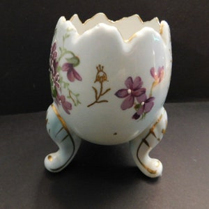 May include: A white porcelain egg-shaped planter with three gold-painted legs. The planter is decorated with purple and green floral designs.