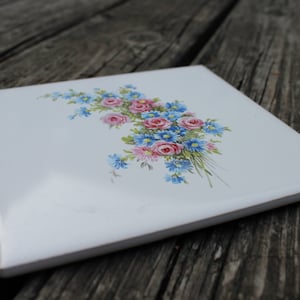 May include: A white ceramic tile with a painted floral design. The design features pink and blue flowers with green leaves.