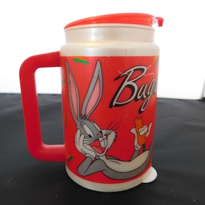 May include: Red plastic Bugs Bunny mug with a white lid and a handle. The mug features a cartoon image of Bugs Bunny with a carrot in his hand. The text "Bugs" is printed on the mug.