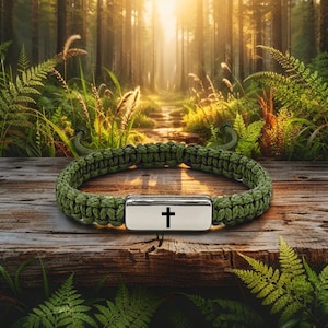 Puede incluir: Una pulsera verde de paracord con un cierre plateado que presenta una cruz negra. La pulsera se muestra sobre una superficie de madera con un fondo de bosque, mostrando un tema natural y al aire libre.