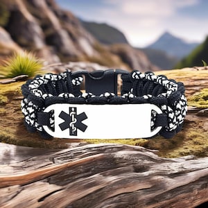 Puede incluir: Pulsera de paracord en blanco y negro con una placa de metal blanco con el símbolo médico de la Estrella de la Vida. La pulsera tiene un cierre de plástico negro y se muestra sobre un trozo de madera.