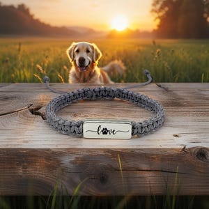 May include: A gray macrame bracelet with a silver rectangular charm. The charm has the word "love" and a paw print design. A golden retriever dog is in the background, with a sunset in the distance.