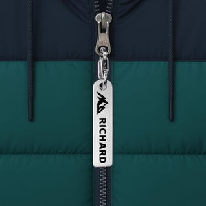 Puede incluir: Un llavero plateado con el nombre "RICHARD" en letras negras, colgado de una cremallera de chaqueta. La chaqueta es azul marino y verde azulado, con capucha y cierre de cremallera.