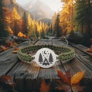 May include: A green paracord bracelet with a silver centerpiece featuring a camping scene with trees, a campfire, moon, and stars. The bracelet is set on a rustic wooden surface with fall foliage in the background.