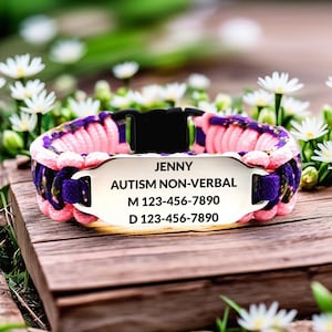 May include: A pink, purple, and black paracord dog collar with a metal nameplate. The nameplate reads "JENNY, AUTISM NON-VERBAL" with two phone numbers. The collar is on a wooden surface with white flowers in the background.