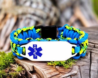 May include: A blue and yellow paracord bracelet with a white metal plate featuring a blue medical symbol. The bracelet has a black clasp and is displayed on a wooden surface with green moss.