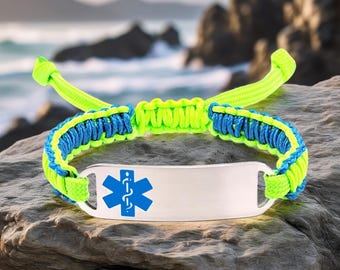 May include: A neon green and blue paracord bracelet with a silver medical ID tag. The tag features a blue medical symbol. The bracelet is resting on a gray rock with a blurred ocean background.