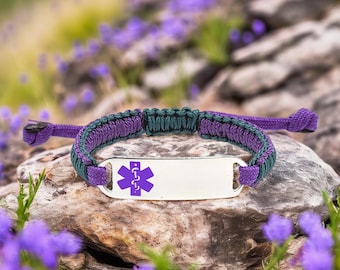 May include: A purple and green paracord bracelet with a silver medical ID tag. The tag features a purple medical symbol. The bracelet is displayed on a rock surrounded by purple flowers.