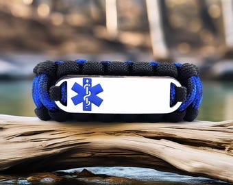May include: A black and blue paracord bracelet with a white metal plate. The plate features a blue Star of Life medical symbol. The bracelet is resting on a piece of driftwood, with a blurred natural background.