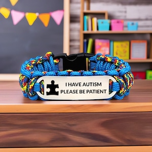 May include: A blue paracord bracelet with a black clasp and a metal plate. The plate has a puzzle piece symbol and the text "I HAVE AUTISM PLEASE BE PATIENT". The bracelet features a colourful woven design.
