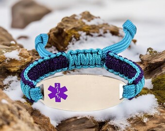 May include: A medical ID bracelet with a silver plate and a purple medical symbol. The bracelet features a woven design with teal and purple cords. The background shows snow and rocks.