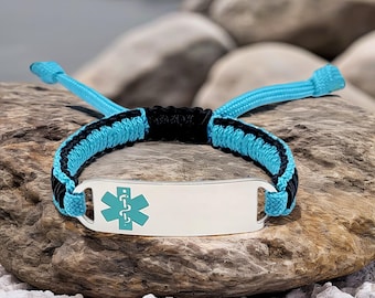 May include: A medical ID bracelet with a silver plate and a turquoise medical symbol. The bracelet features a woven design with black and turquoise cords. The bracelet is displayed on a rock.